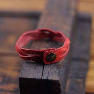 May include: A red braided leather bracelet featuring a circular bronze-toned metal snap closure embossed with a star design. The leather strap includes visible text that reads MADE IN ITALY, and the bracelet is formed by three intertwined leather strands.