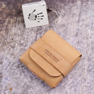 May include: A tan leather wallet with a fold-over flap and a strap closure. The wallet is embossed with the text INEE Be Special HANDCRAFTED LEATHER. A white square tag with a black handprint graphic and text is attached by a thin white string.
