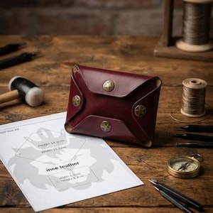 Puede incluir: Una cartera de cuero rojo oscuro con forma de sobre y cuatro botones a presión de color latón con forma de estrella. La cartera se muestra junto a una plantilla de papel con dimensiones de 14,3 cm de alto y 21,1 cm de ancho para el cuero.