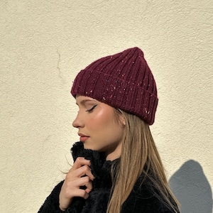Puede incluir: Un gorro de punto burdeos con motas de color. El gorro es acanalado y tiene un borde doblado. La persona de la imagen lleva un abrigo negro y difuso. El fondo es una pared de color beige claro.