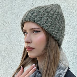 Puede incluir: Un gorro de punto acanalado de color verde apagado. El gorro está hecho de un material suave y texturizado. El gorro se lleva con una bufanda estampada y un abrigo oscuro.