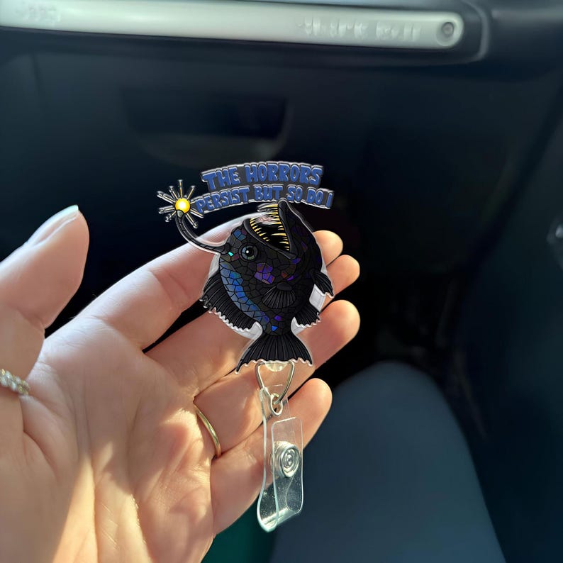 May include: A black anglerfish with a glowing lure, a blue and white text banner that reads "The Horrors Persist But So Do I", and a clear plastic retractable badge holder.