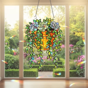 May include: Stained glass hanging succulent planter with a chain. The planter is orange with green and blue succulents and trailing green leaves. The window behind the planter shows a lush green garden.