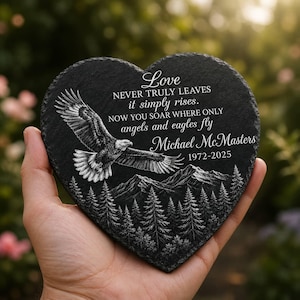 May include: A heart-shaped black slate plaque featuring an engraved eagle soaring above a forest and mountain range. The text reads: Love NEVER TRULY LEAVES it simply rises. NOW YOU SOAR WHERE ONLY angels and eagles fly Michael McMasters 1972-2025.