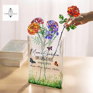 May include: A clear glass vase with a floral design and the text "Mom's Garden Love Grows Here since 2003 Elias Mia Lena Bella". The vase has a green grass design at the bottom and butterflies on the sides.