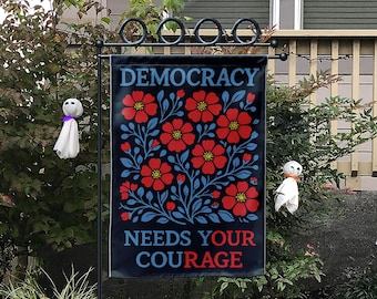 May include: A black garden flag featuring a red floral pattern and the words "DEMOCRACY NEEDS YOUR COURAGE" in white and red. The flag is suspended on a black metal stand. Two small ghost decorations are visible in the background.