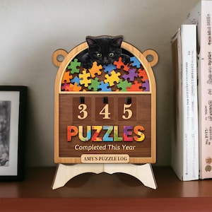 Könnte beinhalten: Holzpuzzle-Logbuch mit einer schwarzen Katze, die über die Oberseite schaut. Das Logbuch zeigt die Zahl "345" und das Wort "PUZZLES" in bunten Buchstaben. Der Text lautet "Completed This Year" und "AMY'S PUZZLE LOG". Die Puzzleteile sind mehrfarbig.