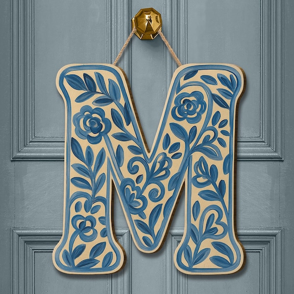 May include: A wooden letter M wall hanging featuring a cream-coloured base with hand-painted blue floral and vine patterns. The letter is suspended by a tan twine cord attached to a gold-toned metal hook at the top, creating a decorative accent.