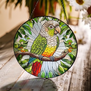 May include: Round stained glass window art featuring a colorful parrot perched on a branch. The parrot has green, yellow, red, and blue feathers. The background includes green leaves and a light blue sky. The window is framed with a dark metal chain.