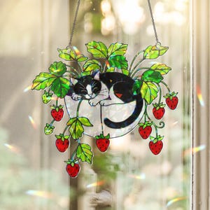 May include: A decorative hanging stained glass piece showing a black and white cat sleeping in a white planter with green leaves and red strawberries. It hangs by a silver chain and is designed to catch the light, creating a rainbow effect.