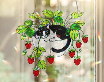 May include: A decorative hanging stained glass piece featuring a black and white cat sleeping in a white planter with green leaves and red strawberries. The piece is suspended by a silver chain and is designed to catch the light.