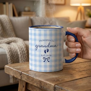 Peut inclure: Mug en céramique à carreaux bleus et blancs, avec une anse et un bord bleus. Le mug porte l'inscription "Promue grand-mère", des empreintes de pas et un nœud, ainsi que l'année "EST. 2026". La tasse contient une boisson chaude.