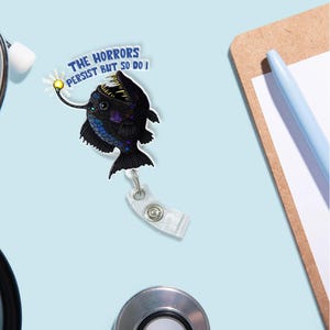 May include: A black anglerfish with a glowing lure, a blue and white text bubble that reads "The horrors persist but so do I", and a clear retractable badge reel.