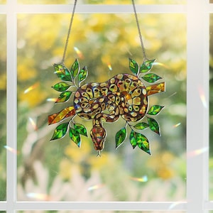 May include: Stained glass suncatcher featuring a snake coiled on a branch with green leaves. The snake has a brown and yellow pattern. The suncatcher hangs from a chain and is set against a blurred outdoor background.