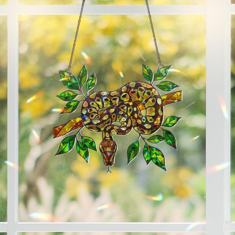 May include: Stained glass suncatcher depicting a snake coiled on a branch with green leaves. The snake has a brown and yellow pattern. The suncatcher hangs from a chain and is set against a blurred outdoor background.