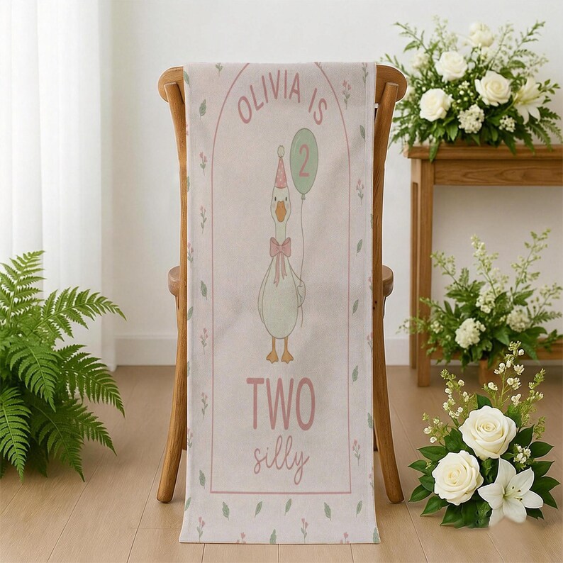 May include: A light pink table runner featuring a cartoon goose holding a balloon. The runner has the text "Olivia is 2" and "Two silly" in pink script. Floral and leaf accents border the design.
