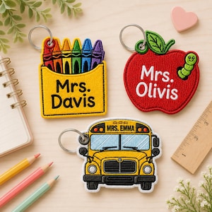 May include: Three embroidered keychains with metal rings. One is a yellow crayon box labelled Mrs. Davis, one is a red apple with a green worm labelled Mrs. Olivis, and one is a yellow school bus labelled Mrs. Emma, shown with a ruler for scale.