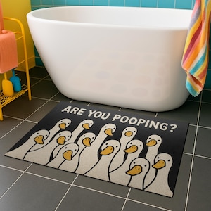 Puede incluir: Alfombra de baño rectangular negra con un patrón repetido de gansos blancos de dibujos animados con picos amarillos. El texto "ARE YOU POOPING?" está impreso en blanco. La alfombra está colocada frente a una bañera blanca.