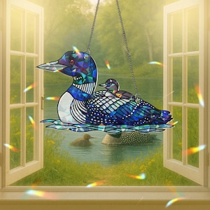 Puede incluir: Un vitral colgante con un colimbo azul e iridiscente con un colimbo más pequeño en su espalda. El colimbo está en un entorno de lago con vegetación verde y un cielo azul.