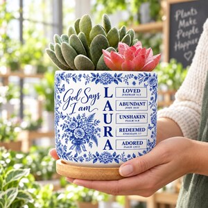 May include: A white ceramic planter with a blue floral pattern and the text God Says I am LAURA followed by the words LOVED, ABUNDANT, UNSHAKEN, REDEEMED, and ADORED with corresponding Bible verses. The planter sits on a round bamboo saucer.