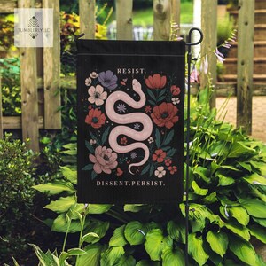 Könnte beinhalten: Schwarze Gartenflagge mit einem rosa Schlangenmuster, umgeben von roten, rosa und lila Blumen. Die Worte "RESIST" und "DISSENT.PERSIST." sind in Weiß gedruckt. Die Flagge wird im Garten gezeigt.