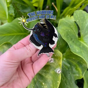 May include: A black anglerfish with a glowing lure on a white background with the text "The Horrors Persist But So Do I". The anglerfish is on a retractable badge reel.
