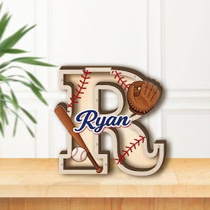 Könnte beinhalten: Ein mehrschichtiger Holzbuchstabe R im Baseball-Design mit einem braunen Schläger, einem weißen Baseball und einem braunen Lederhandschuh. Der Buchstabe hat rote Baseball-Nahtmuster und den Namen Ryan in blauer Schrift in der Mitte.