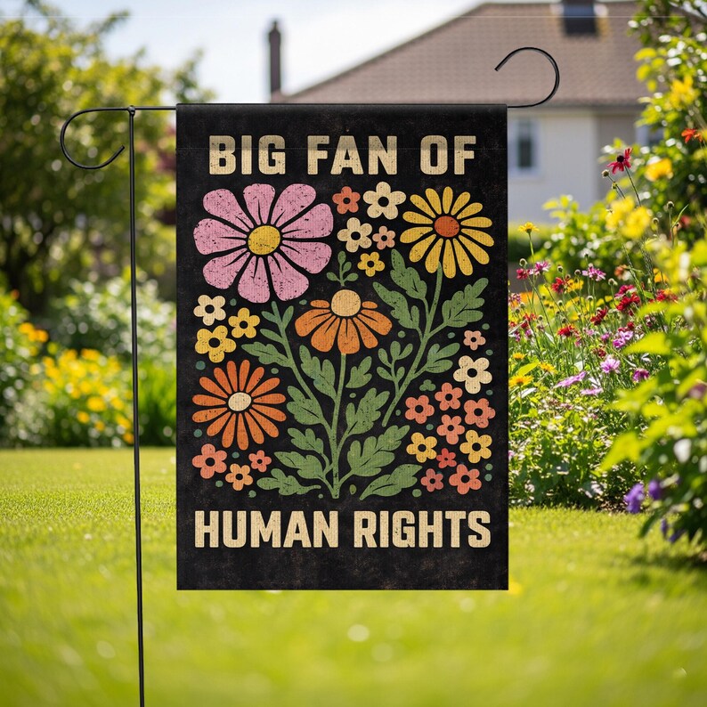 May include: A rectangular garden flag with a black background featuring the text BIG FAN OF HUMAN RIGHTS in a bold, light-colored font. The design includes a central arrangement of stylized flowers in shades of pink, yellow, and orange with green leaves.