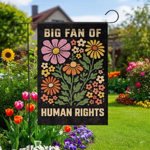 May include: A rectangular black garden flag featuring the text BIG FAN OF HUMAN RIGHTS in a bold, light-colored font. The design includes a central illustration of various stylized flowers in shades of pink, yellow, and orange with green leaves.