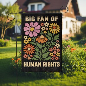 May include: A rectangular black garden flag featuring the text BIG FAN OF HUMAN RIGHTS in a bold, light-colored font. The center displays a colorful illustration of various flowers in shades of pink, yellow, orange, and white with green stems.