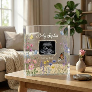 May include: A clear acrylic block with floral and butterfly accents, featuring the text "Baby Sophia" and an ultrasound image. The text "Arriving 2026" is also visible. The block is displayed on a wooden table.