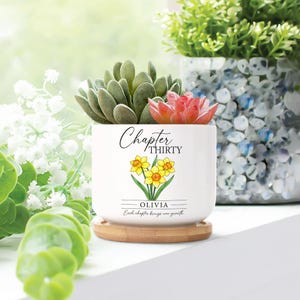 May include: A white ceramic planter with the text "Chapter Thirty" and a daffodil illustration. The planter holds a succulent and a small red flower. It sits on a round wooden coaster. The text "Olivia" and "Each chapter brings new growth" are also on the planter.