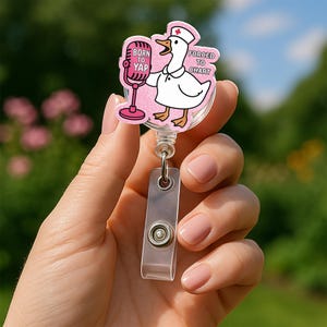 May include: A pink and white cartoon goose wearing a nurse's hat and holding a microphone. The goose is singing into the microphone and the text "Born to Yap" is written on the microphone. The text "Forged to Chart" is written on the goose's right side. The goose is attached to a clear plastic retractable badge holder.