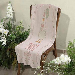 May include: A pale pink table runner with a cartoon goose holding a balloon with the number 2, a pink bow, and the words "TWO silly". The runner is draped over a wooden chair. Green and pink floral accents are also present.