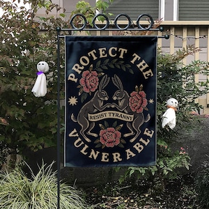 Könnte beinhalten: Eine rechteckige Gartenflagge mit schwarzem Hintergrund, die zwei einander gegenüberstehende Hasen zeigt, umgeben von rosa Blumen und grünen Blättern. Der Text lautet PROTECT THE VULNERABLE in einem Bogen und RESIST TYRANNY auf einem Banner.