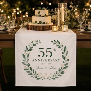 May include: A white fabric table runner featuring a printed green botanical wreath design. The center text reads 55th ANNIVERSARY EST. 1969 and Jessie & Adam. The design includes green leaves and gold accents, centered on a rectangular textile.