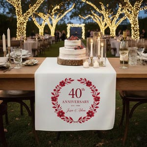 May include: A white table runner with a red floral wreath design, celebrating a 40th anniversary. The text includes "40th Anniversary" and the names "Jessie & Adam". A two-tiered cake sits on a wooden base.