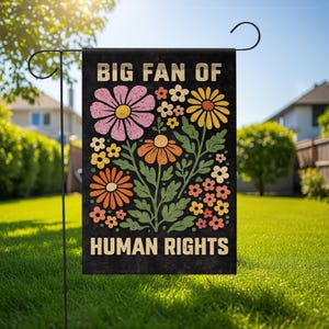 May include: A rectangular garden flag with a black background featuring the text BIG FAN OF HUMAN RIGHTS in a bold, light-colored font. The design includes a central illustration of various colorful flowers in pink, yellow, and orange.