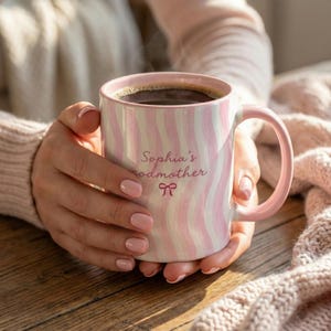 Puede incluir: Taza de café a rayas rosas y blancas con asa y borde rosas. La taza está llena de café y presenta el texto "Sophia's godmother" con un lazo. La taza está sostenida por una persona.