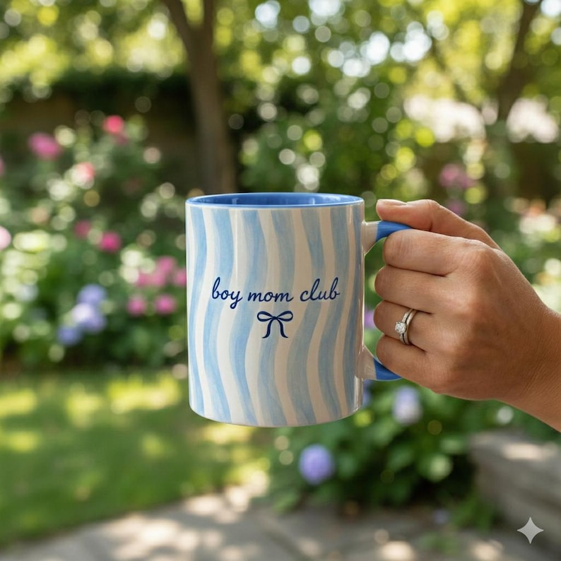 May include: A white ceramic mug with blue wavy stripes and a blue interior and handle. The mug has the text "boy mom club" and a bow graphic in blue. The mug is held in a hand against a blurred outdoor background.