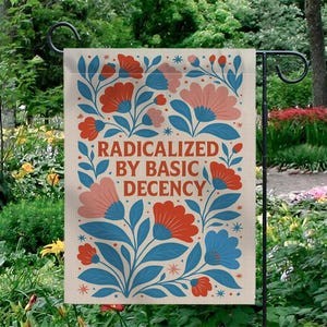 Op de afbeelding: Een tuinvaandel met een bloemenontwerp in rood, blauw en roze. De vlag heeft de tekst "RADICALIZED BY BASIC DECENCY" in een gestileerd lettertype. De vlag hangt aan een zwarte metalen standaard in een tuin.