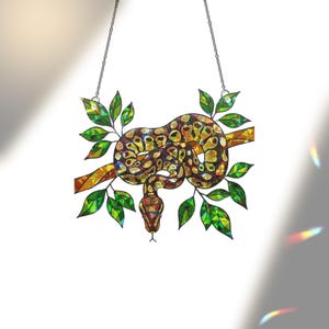 May include: Stained glass snake suncatcher. The snake has a brown and yellow patterned body, coiled around a branch with green leaves. The snake's head is red, and the piece hangs from a chain.