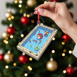 May include: A rectangular Christmas ornament depicting a swimming pool scene with Santa Claus in a floatie. The ornament features a ladder, beach ball, candy canes, and other pool toys. The ornament is held by a red ribbon against a blurred Christmas tree background.