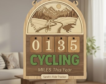 May include: Wooden cycling miles tracker with a mountain and cyclist design. The counter shows "0135" miles. The text "CYCLING MILES This Year" is displayed in green. The tracker is personalized with "Sarah's Ride Tracker".
