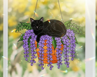 Puede incluir: Adorno de ventana de vidriera con un gato negro sobre una maceta de glicinas moradas. La maceta naranja tiene un diseño geométrico. Hojas verdes rodean las flores y el gato. Colgando de una cadena.