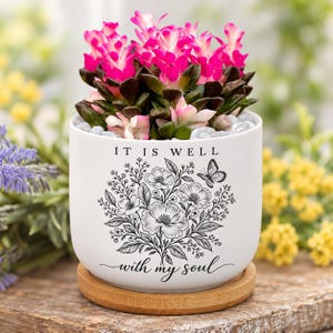 Peut inclure: Un pot de fleurs en céramique blanche avec une soucoupe en bambou contenant une plante succulente aux feuilles rose vif et vert foncé. Le pot présente une illustration au trait noir d'un bouquet floral, un papillon et le texte IT IS WELL with my soul.