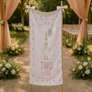 May include: A linen banner with the text "Olivia is 2" and "Two silly" in pink. The banner features a cartoon goose holding a balloon. The banner is decorated with floral accents and tied with bows.