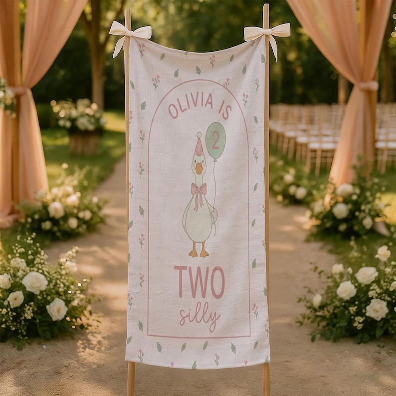 May include: A linen banner with the text "Olivia is 2" and "Two silly" in pink. The banner features a cartoon goose holding a balloon. The banner is decorated with floral accents and tied with bows.
