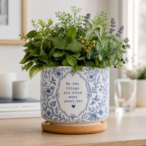 May include: A white ceramic planter with a blue floral and bird pattern, featuring the text "Be the things you loved most about her" inside a decorative frame. The planter sits on a round wooden saucer and contains a lush arrangement of green foliage.