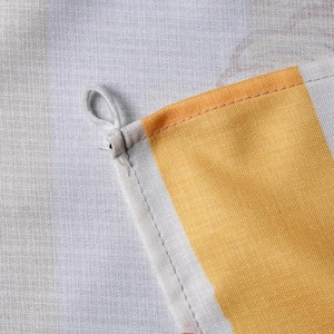 Puede incluir: Un pa&ntilde;o de cocina de tela tejida con un patr&oacute;n de cuadros blancos y gris claro con una prominente franja vertical amarilla. La esquina del pa&ntilde;o incluye un peque&ntilde;o lazo de tela blanca para colgar, con costuras visibles a lo largo del dobladillo.
